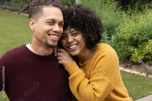 African American couple sitting close in garden wearing knit sweaters, leaning and showing ring