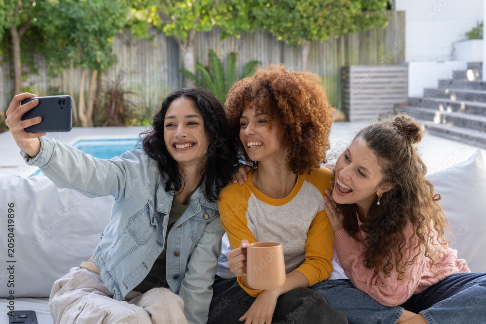 Obraz premium Diverse female friends taking selfie on light sofa by pool, holding smartphone and ceramic mug