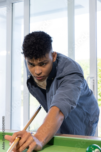 African American teen leaning over pool table at home, aiming wooden cue in dark-blue overshirt