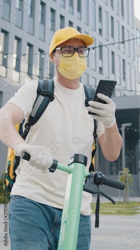 A delivery person wearing a mask and gloves on an electric scooter, holding a phone, in a vertical shot.