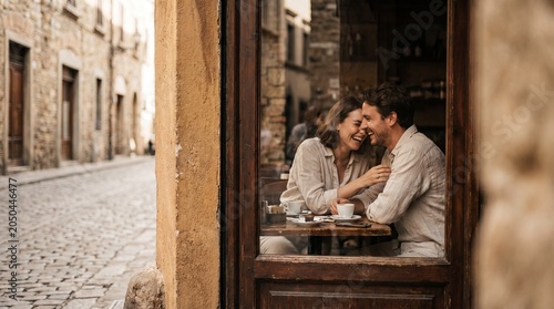 A joyous young couple sharing a laugh while sitting at a table with coffee inside a cozy cafe, seen through a window in a charming European cobblestone street.