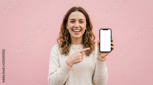 A smiling woman points to a smartphone screen against a pink background. She is wearing a white sweater and appears happy and confident.