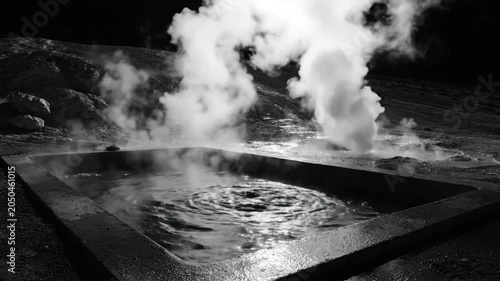 Black and white shot of a steaming hot spring pool with swirling water and rising vapor on a dark landscape