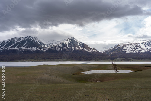 Panoramic View of Alpine Lake with Snow-Capped Mountain Range and Lone Tree Under Dramatic Storm Clouds, Mongolian Altai