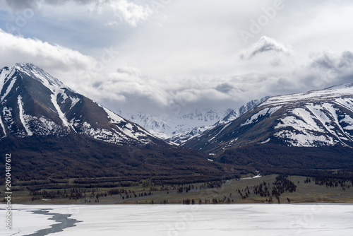 Frozen Alpine Lake with Melting Ice, Larch Forest and Snow-Capped Mountain Range Under Dramatic Clouds, Mongolian Altai