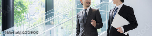 新緑のオフィスの窓際で会話する若手ビジネスマンと上司、取引先のコミュニケーション商談や指導ojtなどの仕事イメージの上半身顔無しのバナー