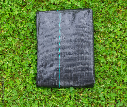 Folded black weed control fabric placed on a green grassy lawn. Durable woven landscape fabric used for weed suppression, soil protection, gardening, horticulture, and landscaping projects
