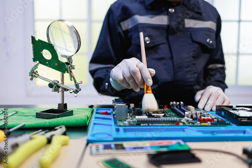 Technician in work uniform cleaning a computer motherboard with a brush at an electronics repair desk with tools, magnifier and parts in a bright workshop.