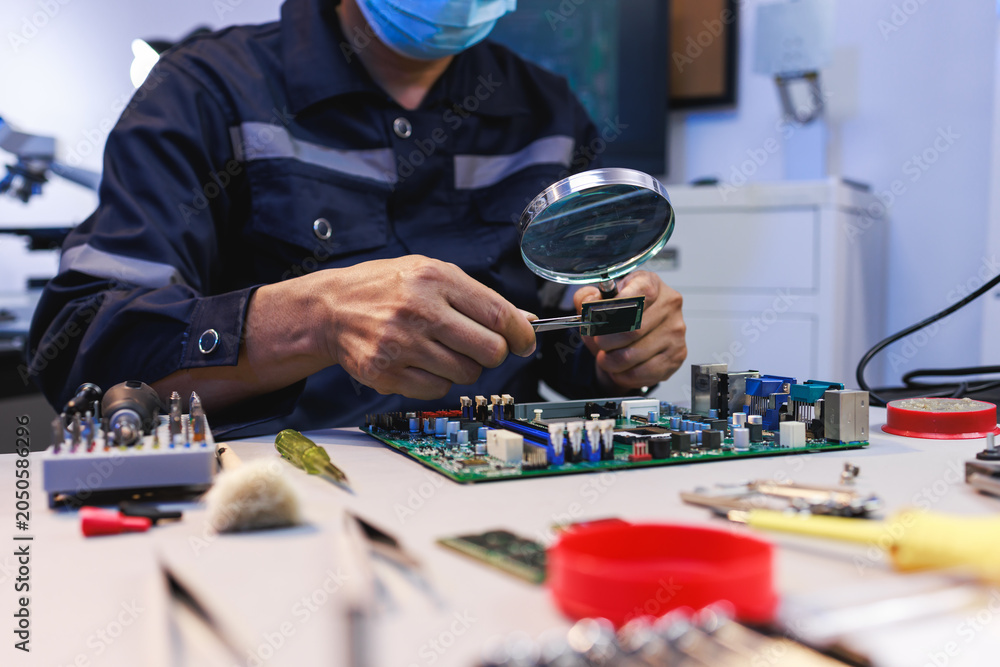 Obraz premium Technician inspecting a circuit board with a magnifying glass at an electronics repair workbench surrounded by tools and components.