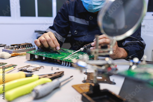 Fotografie Close view of a masked technician repairing a green circuit board at a workbench with tools and magnifier in an electronics workshop