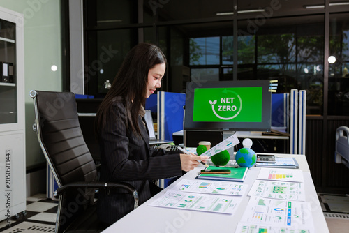 Businesswoman reviewing sustainability reports at an office desk with a net zero screen, charts and eco models, showing climate planning, green business goals and clean energy strategy.