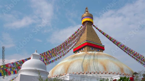 A static medium shot of the grand Boudhanath Stupa featuring a smaller white dome in the foreground, decorated with colorful prayer flags under a bright sky in Nepal.