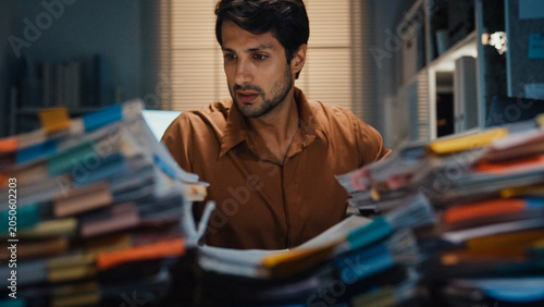 Overworked young Latin businessman working late on document in large stack of paperwork files at office night. Tax audit deadline, accounting stress, corporate burnout, urgent business concept.