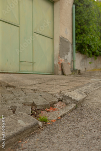 Low-angle view of a cracked concrete driveway edge with loose paving slabs and small weeds growing between them, leading to a pale green metal garage door set