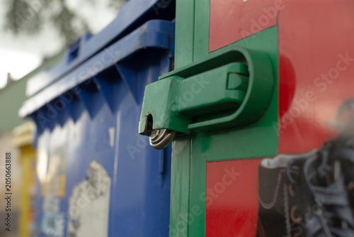 Shallow depth-of-field close-up showing a green metal latch secured on the side of a red recycling container. A blue plastic waste bin and other out-of-focus