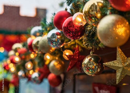 Schilderij op canvas Bright red, gold, and silver Christmas ornaments hang on green garland, illuminated by warm lights