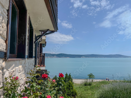 Landscape view of a traditional hotel building facade with red roses blooming in the yard and a panoramic view of a calm turquoise sea bay under a blue sky