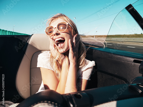Portrait of young beautiful and smiling hipster female in convertible car. Sexy carefree woman driving cabriolet. Positive model riding and having fun in sunglasses at sunset
