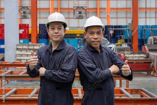 Fotografia Two worker in a hardhat and mechanic coverall dust protection suit stand confidently, giving a thumbs-up and smiling in the bustling industrial environment