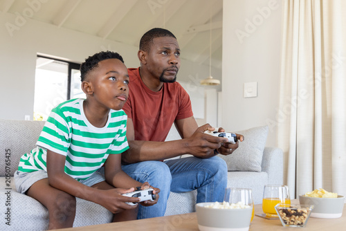 African American dad and child playing console games at home on sofa with gamepads, popcorn, juice