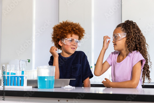 Two children wearing safety goggles mixing blue liquid in test-tube rack at school bench