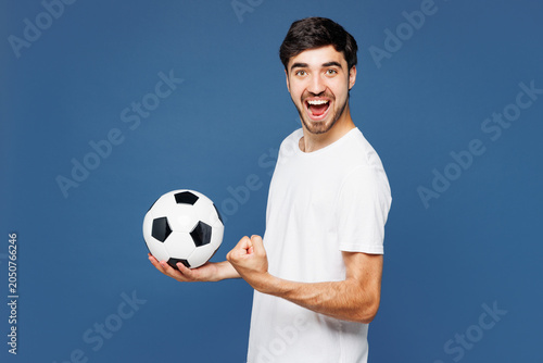 Young man fan wears basic white t-shirt cheer up support football sport team hold in hand soccer ball watch tv live stream doing winner gesture celebrate clench fist isolated on plain blue background.