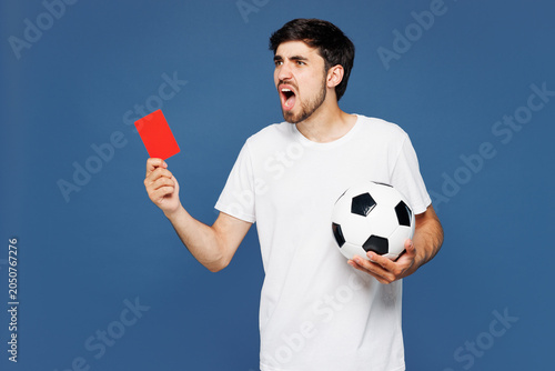 Young referee man fan wear basic white t-shirt cheer up support football sport team hold soccer ball, red card propose player retire from field watch tv live stream isolated on plain blue background.