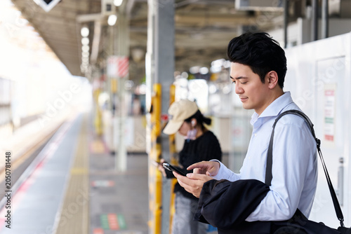 Photos 駅のホームでスマートフォンを操作する30代の日本人男性会社員と背景にいる女性