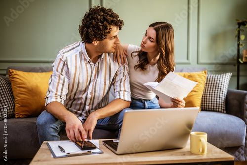 A young couple sits together in their living room, reviewing paperwork and discussing important financial matters.