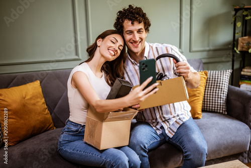 Adult couple is unboxing their packages delivered to home address. They are taking selfie with new items.