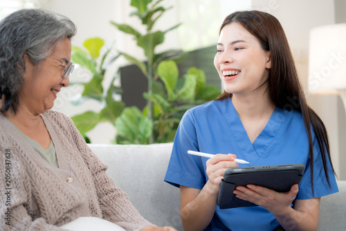 Asian woman caregiver using digital tablet for senior patient consultation, professional nurse checking health record in living room at home, elderly care assessment with checkup, health care support.