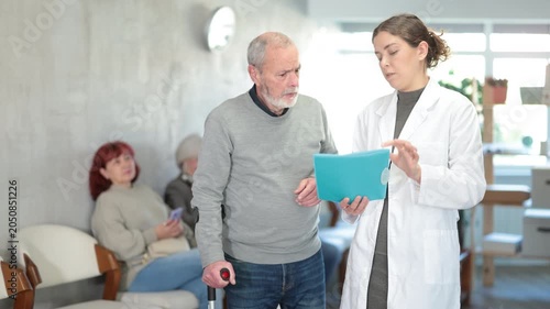 Positive polite young nurse or doctor talking to aged man patient in reception area in background queue of people