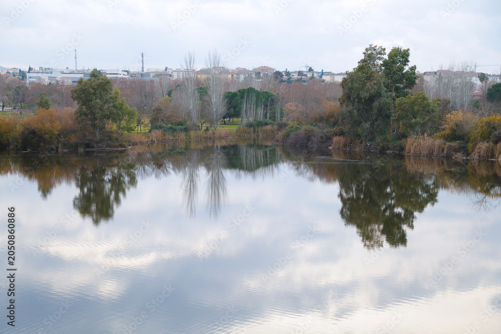 Obraz premium Scenic view of the Guadiana River with perfectly calm water creating symmetrical tree and cloud reflections next to the Guadiana River Park in Merida, Extremadura, Spain.