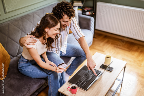Adult couple is sitting at sofa at home and using laptop. They are shopping online. Woman is holding credit card.