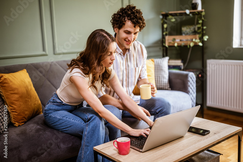 A smiling couple spends quality time at home, exploring content on a laptop. Their relaxed expressions and casual setting suggest online shopping or financial planning.