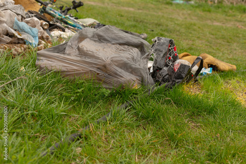 A pile of discarded belongings and rubbish lies on a green grassy field, including a crumpled black plastic bag, a patterned backpack, scattered clothing