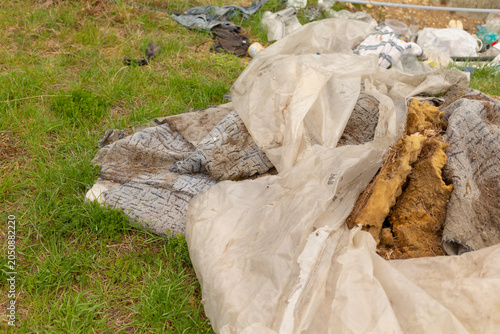 A pile of discarded building insulation, torn fabric and plastic sheeting dumped on a patch of green grass. The scene shows weathered fibrous insulation