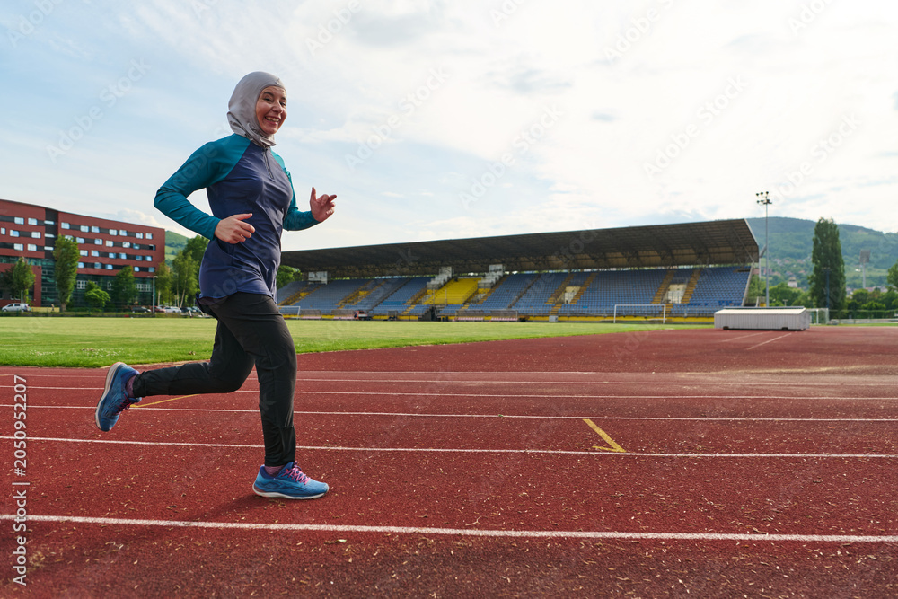 Obraz premium Smiling Muslim Woman Running on Track in Hijab Sportswear at Outdoor Stadium for Fitness