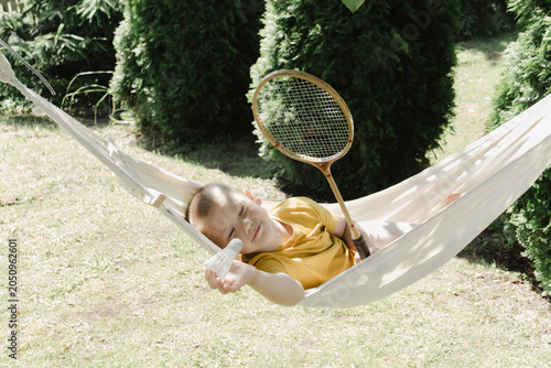 Schilderij op canvas A cute little blond-haired smiling boy of European appearance in a yellow shirt is relaxing and having fun in a light hammock against the backdrop summer green garden