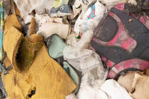 Close-up view of a worn pink and black backpack, shredded yellow car upholstery and assorted trash piled together in a landfill environment. The scene shows