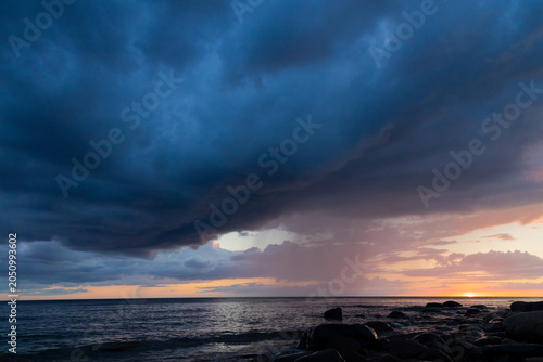 Dramatic Storm Over Ocean At Sunset With Rocky Shoreline And Rainfall On Horizon