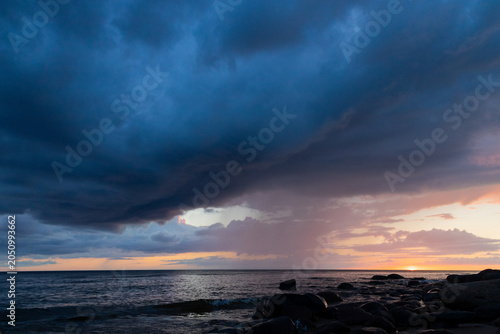 Dramatic Storm Over Ocean At Sunset With Rocky Shoreline And Rainfall On Horizon
