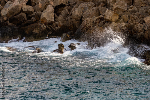 Sea water wave splashing with white foam and spray on rough rocky cliff shore in sunny daylight in December. Coastal nature view, Cape Greco, Ayia Napa, Cyprus.