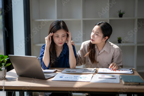 Coworker comforting stressed asian woman with headache at office desk