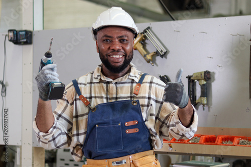 Photos Portrait of happy African carpenter wearing white helmet and apron, holding an electric drill and giving thump up