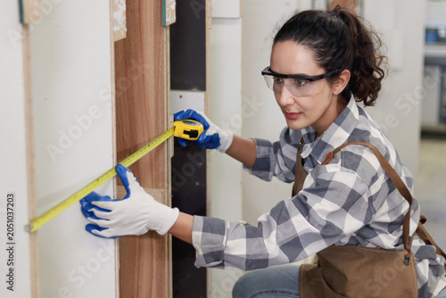 Young woman carpenter wearing safety glasses goggles and apron, holding tape measure for measuring wood plank. Beautiful happy female craft worker creating wooden furniture in carpentry workshop.