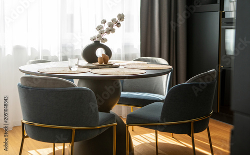 Modern dining room interior with a round wooden table, dark blue velvet chairs with gold legs, and a stylish ceramic vase with cotton flowers near a bright window. Real estate concept