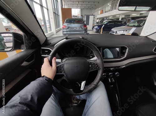 Person sitting in new car at dealership showroom holding steering wheel, first person view
