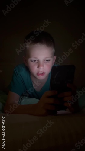 Vertical. A child holds a smartphone under the blanket's light, scrolling through content. The boy uses gadgets while getting ready for bed, his face illuminated by the blue light of the smartphone sc