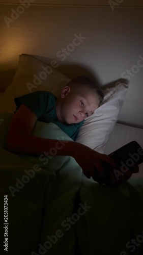 Vertical. A child holds a smartphone under the blanket's light, scrolling through content. The boy uses gadgets while getting ready for bed, his face illuminated by the blue light of the smartphone sc